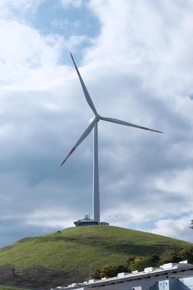 A photo up a green grass hill where ontop next to a solar roofed Pavillon a large wind turbine stands. A few birds are flying around the Pavillon, clouds are hanging scattered in the sky. It’s almost a solar punk esk image