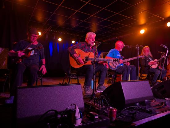 Fairport Convention sitting on chairs on a stage. Ric Sanders is on the left wearing a beanie and a tshirt and holding a violin bow, Simon is next to him holding a guitar and speaking, Peggy is next to him in a bright blue t-shirt and Chris is on the right, suit jacket, long hair, mandolin
