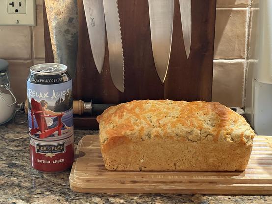 A kitchen countertop. On the left is a colourful empty can of beer. On the right is a loaf of cheese/beer bread on a wooden board. The loaf has plenty of melted cheese on top. There are kitchen knives hanging from a magnetic holder against the wall behind the bread.