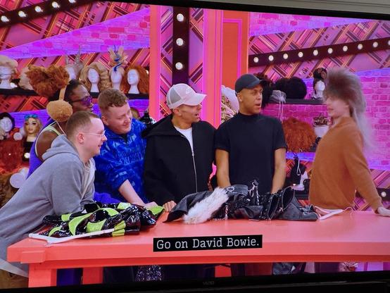 In the Drag Race UK werk room, a group of the queens, dressed in boy mode, observe Viola at the right wearing a heavily teased semi-mullet of a wig; one of the other queens says "Go on, David Bowie."