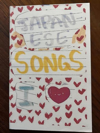 zine cover - title is japanese songs i heart. all writing is done with color markers. background is small red hand drawn hearts, like wallpaper, somewhat evenly spaced and sized. over that is a collage of words written on lined paper. at the top in light purple or gray, the word japanese. then in yellow the word songs. then the word i in light blue. and finally, a smiling red heart, with two tiny white eyes and a small white smile between them, all nearly level.