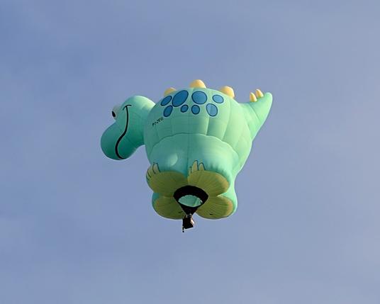 An adorable special shape hot air balloon in the shape of a smiling dinosaur. it's mainly aqua with blue spots and yellow back and tail spikes.