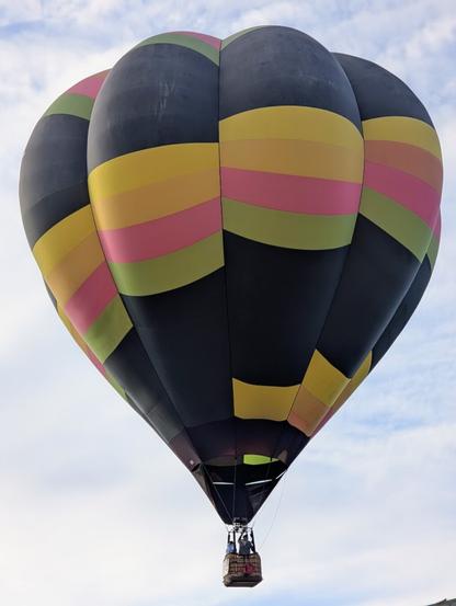 A hot air balloon drifting across a cloudy sky; it's primarily black with horizontal yellow, pink, and green stripes.