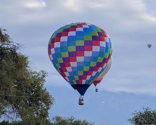 A beautiful multicolored balloon drifting across a stormy sky. There are trees to the left and below it along with a few other balloons in the background.
