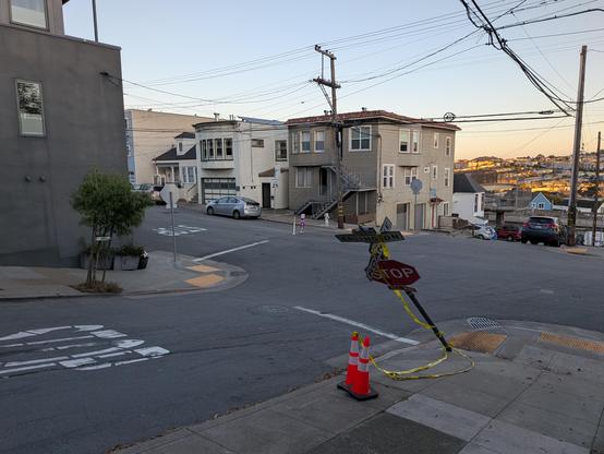 A stop sign on a sidewalk, vaguely draped with caution tape and a couple of traffic cones.  It's bent over at about a 45° angle, and given the steep grade of the street it's facing, that brings it fairly fairly close to the ground.  The street on the opposite side of the intersection is a dead end.  The direction the sign post was hit from is indicated by a freshly-cut arc gouged out of the asphalt coming from one of the cross streets (which also has stop signs, and forms one end of a Slow Street.)