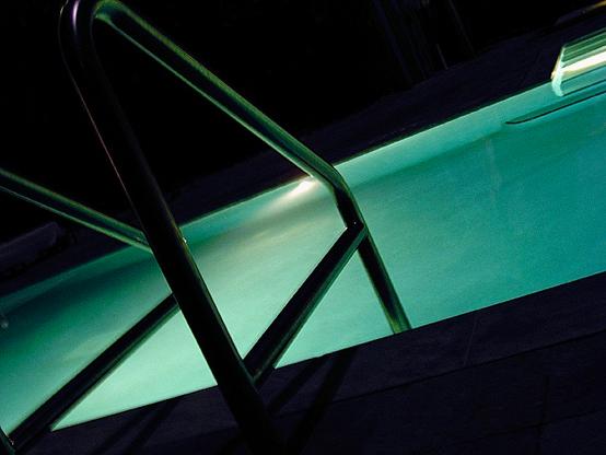 View of a swimming pool at night. The lights are on in the pool, so the water is glowing an aqua color. There is a silhouette of the aluminum rail and stair to use when climbing out of the pool. The rest of the image is black.