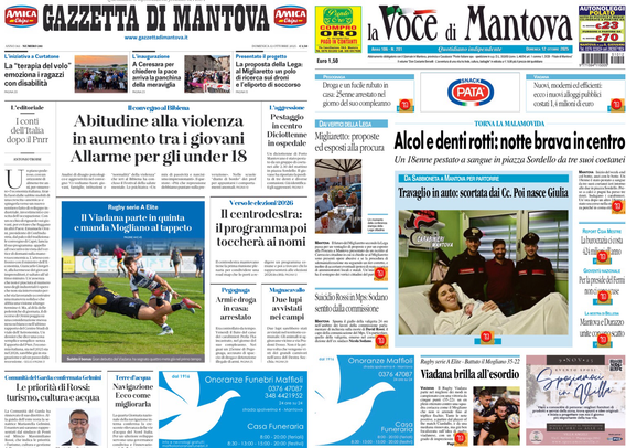Pagina di giornale visualizzata su uno schermo in un ambiente buio. Sono visibili le prime pagine di due quotidiani locali: "Gazzetta di Mantova" a sinistra e "La Voce di Mantova" a destra. I titoli principali parlano di temi sociali come la violenza tra i giovani e di un episodio di bravata in centro collegata ad alcol e dentifrici. Ci sono anche immagini di persone e notizie sportive in evidenza, con una foto di una partita di rugby nella prima pagina a sinistra e un gruppo di giovani sorridenti nella pagina a destra. Nella parte inferiore delle pagine spiccano annunci funebri su sfondo azzurro.