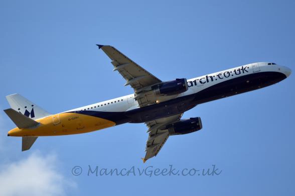 Side view of a twin engined jet airliner flying from left to right at a low altitude, with undercarriage stowed, flaps extended behind the wings, and the nose raised sharply, suggesting it has just taken off.
The plane is mostly white, with a deep purple belly the sweeps up toward the top of the rear fuselage, bright yellow rear fuselage, and deep purple web address "monarch.co.uk" on the forward fuselage, covering the forward cabin windows.
The black registration "G-OZBU" is on the lower rear fuselage, with a stylised black crown that more than resembles an upper case letter "M" on the white tail.
Blue sky fills the rest of the frame, with a fluff of white cloud in the bottom left corner.
