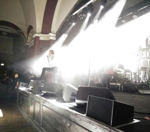 Foto von einer Bühne, auf der gerade eine Band spielt. Die Bühnenscheinwerfer sind nach unten-vorn gerichtet und strahlen krass weißes/helles Licht ab, sodass fast alles auf der Bühne überlichtet ist, außer der Sänger, welcher vor "der Lichtwand" steht und ins Mikro singt, mit einem Bein auf der Monitorbox.