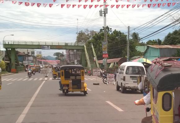 A busy street scene featuring several colorful tricycles, a white van, and motorcycles. There is a pedestrian overpass in the background, with festive red banners hung across the street. Shops and greenery are visible along the sides of the road.

Eine geschäftige Straßenszene mit mehreren bunten Dreirädern, einem weißen Lieferwagen und Motorrädern. Im Hintergrund befindet sich eine Fußgängerüberführung mit festlichen roten Bannern auf der anderen Straßenseite. Geschäfte und Grün sind an den Straßenranden sichtbar.
