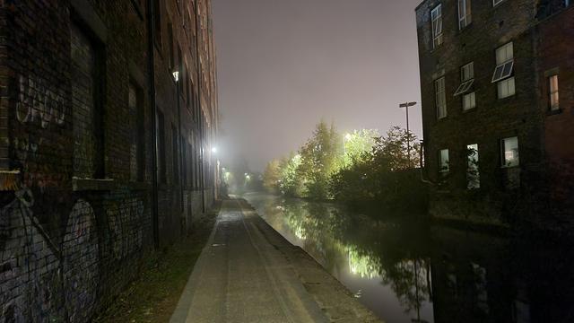 Looking over a more industrial section of the Ashton canal. The pathway is lit by white lighting, and to the right runs the canal. The water reflects some foliage in the distance, and closer, Theo old industrial buildings flank the canal.