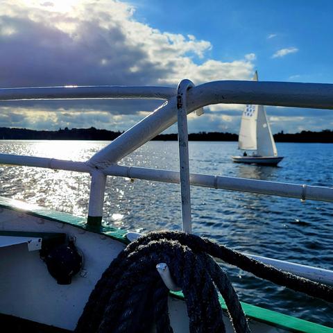 Blick auf eine Reling mit Tauwerk vor Gewässerkulisse, blauem Himmel mit Wolken, durch die die Sonne blitzt und sich im wogenden Wasser widerspiegelt. Rechts im Bildhintergrund unscharf ein Segelboot, recht nah am Schiff, von dem aus fotografiert wird.