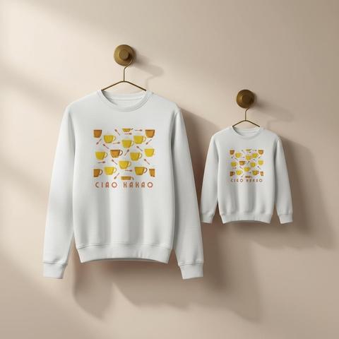 Zwei weiße Sweatshirts im Partnerlook hängen nebeneinander an minimalistischen Wandhaken. Das größere Modell und die Kinderversion zeigen das warme Retro-Design „Ciao Kakao“ mit senfgelben und braunen Kakaotassen, weißen Sahnehäubchen und kleinen Löffeln. Der Druck wirkt lebendig auf dem klaren Weiß und verbreitet gemütliche, moderne Vintage-Atmosphäre.