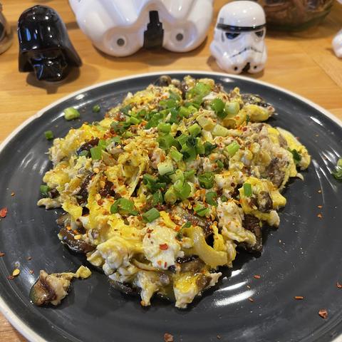 Na zdjęciu jajecznica z kawałkami leśnych grzybów, posypana szczypiorkiem i płatkami chili, podana na ciemnym talerzu. W tle figurki z uniwersum Star Wars