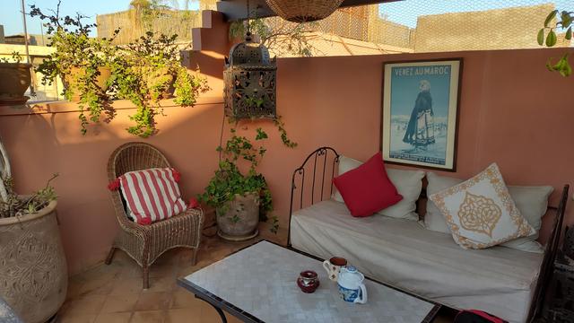 Dachterrasse. Rote Wand vor der ein Sofa aus Metall steht mit weißen Polstern und farbigen Kissen. Grüne Pflanzen auf der Mauer. Ein Großteil ist im Schatten, von links oben kommen Sonnenstrahlen. Sieht gemütlich und entspannt aus