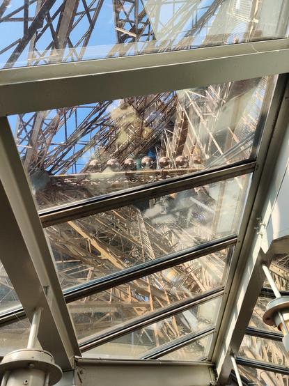 Blick durch eine Glasscheibe von innen auf eine Stahlkonstruktion (die des Eiffelturms) durch die der blaue Himmel scheint.