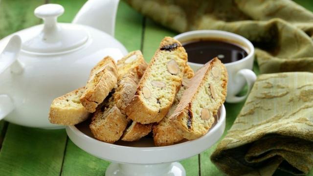 Biscotti con almendras y pasas sobre una fuente alta blanca, al lado de una taza de café y una tetera blanca, sobre una mesa verde.