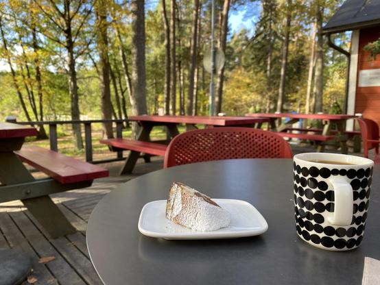 Kahvilan terassi, lähimpänä pyöreällä pöydällä aloittamaton kakkupala ja kahvikuppi. Taempana puisia pöytä- penkki-yhdistelmiä. Ihmisiä ei ole. Taustalla jykevää rantametsikköä, mäntyjä, koivuja ja leppiä. Koivun lehdet kellastuneet.