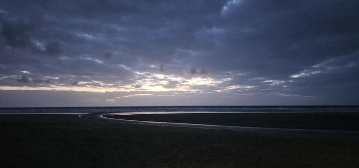 A beach at dusk, with pale light shining off a curving river that cuts a crescent along the sand on its way to the waves. The fading light highlights the heavy grey and white clouds.