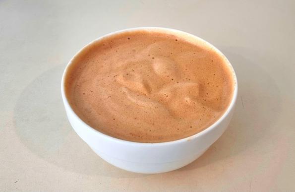 A flat, wide white porcelain bowl containing a hot beverage that cold l fills out almost to the brim. The top of the drink has a visible thick layer of brown-orange foam.