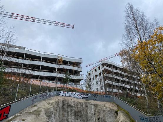 Kaksi rakenteilla olevaa kerrostaloa, niiden yllä nosturit. Parvekkeiden reunat aaltoilevat sisään ja ulos.