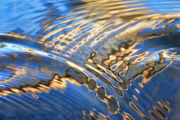 Abschnitt einer Spiegelung auf der Oberfläche eines kleinen Bachlaufes. Die Gold- und Blautöne werden durch kleine Wellen unterbrochen