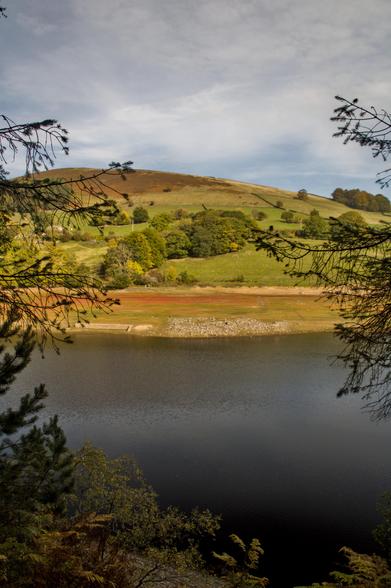 A deep blue reservoir with ruins at the water's edge, with rolling green farmland in the background and rocky edge in the distance