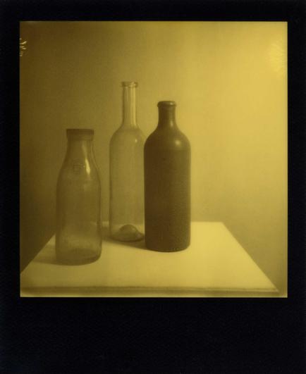 Zwei Glasflaschen und eine dunkle Tonflasche stehen auf einem weißen Sockel vor einer grauen Wand. Die linke Flasche ist unter dem Flaschenhals mit einem großen "G" gekennzeichnet. Das Foto ist in Grau- und Gelbtönen gehalten, weil die Aufnahme mit einem Yellow-Film aufgenommen wurde. Das Foto ist von einem schwarzen Bildrand umgeben.
