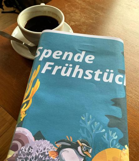 Bild zeigt Tasse mit Kaffee auf dem Tisch, davor Spendendose mit dem nautischen Motiv und der Schrift "Spende Frühstück"