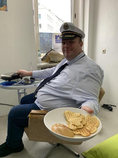 Bild zeigt AgentK im Kapitänskleisund, auf einem Sessel, der Waffeln anreicht und im Hintergrund das Waffeleisen....