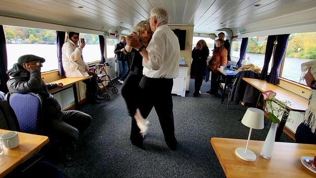 An Bord eines Schiffes unter Deck: ein tanzendes Paar in Bildmitte. Drumherum schauen interessierte Personen stehend und sitzend dem Tanzpaar zu. Ihre Gesichter sind unkenntlich gemacht. Fenster zur linken und rechten Bildseite geben den Blick auf ein Gewässer frei.
