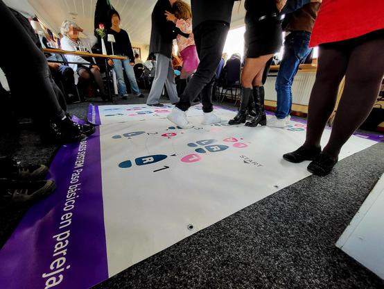 An Bord eines Schiffes unter Deck: Mehrere Personen versuchen sich in die Schritte des Tangotanzes. Man sieht viele Beine. Gesichter sind unkenntlich gemacht. Am Boden eine Plane, auf der Fußschritte in blau und rosa markiert sind mit Buchstaben "R" für rechts und "L" für links.