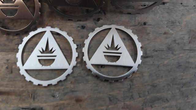 2 Metaltokens, rund, umfasst von einem Zahnkranz, in der Mitte ein Dreieck aus dem das Symbol eines Kessels mit dem Symbol einer Explosion darüber dargestellt ist.