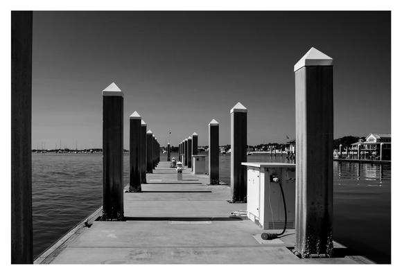 Black and white image of a serene marina dock with evenly spaced tall pillars. Calm water and distant boats under a clear sky in the background.