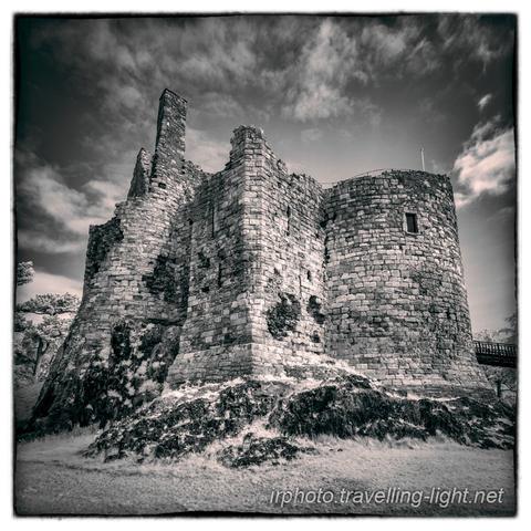 A toned black and white infrared photo looking up at a ruined castle on a small rocky hill.