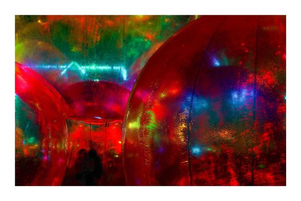Colorful abstract scene with large, shiny, reflective spheres in shades of red, green, and blue, creating a vibrant and immersive environment. Texture on the spheres adds a shimmering effect. Two silhouetted figures are visible in the lower left corner, suggesting interaction or movement within the space.