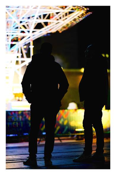 Silhouettes of two people standing in front of a brightly lit structure at night. The background features colorful barricades and blurred lights, creating a vibrant atmosphere.