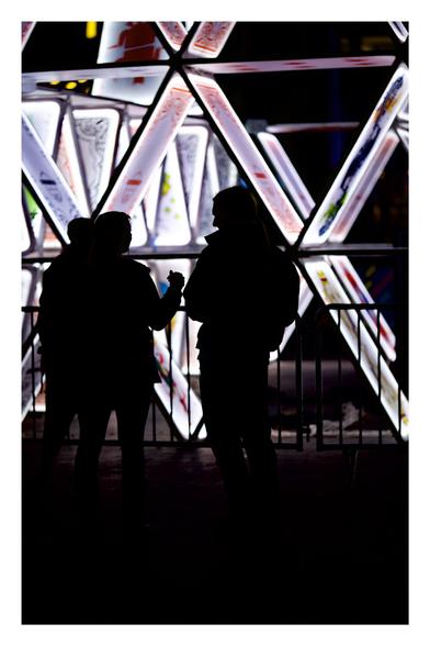 Silhouettes of two people in front of a large, illuminated playing card structure. The cards are arranged in a triangular formation with bright light highlighting their edges. A metal barrier separates the people from the display, emphasizing the glowing cards in the background.