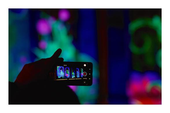 A hand holds a smartphone horizontally, capturing bright, colorful neon abstract shapes in the background. The phone's screen displays a live view of the vibrant scene with detailed hues of blue, green, and pink. The setting appears dark, emphasizing the illuminated colors on both the phone and the backdrop.