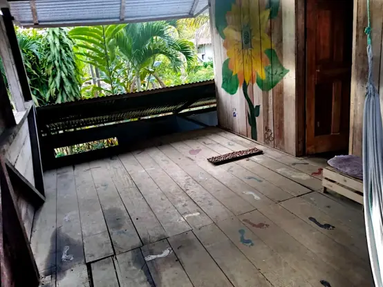 The front room of the loft in the rustic wooden house features excellent airflow due to being open on the northwest side. Ornamental plants grow up against the lower part of the roof, and the other house is visible in the distance. The walls and floor of the room are adorned with hand- and foot-painted art including a large sunflower mural. A hammock hangs in the right of the photo.