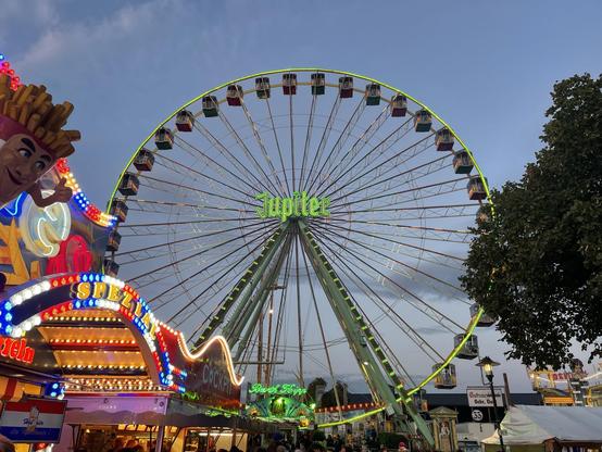Mittiger Blick auf ein Riesenrad auf dem Dürkheimer Wurstmarkt. Das Risenrad hat viele Gondeln, ist grün beleuchtet, und in der Mitte steht der Name Jupiter in Leuchtschrift.