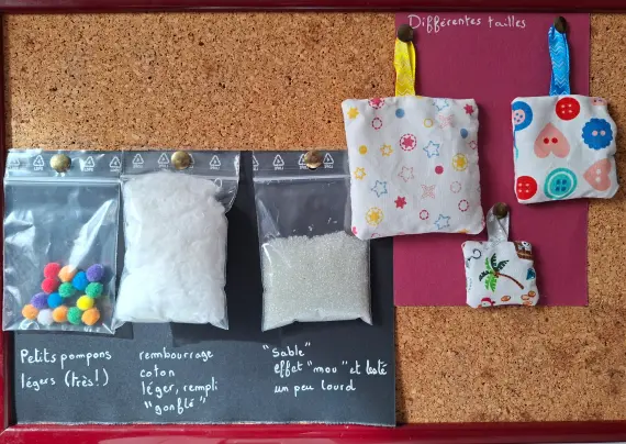 Tableau en liège où sont accrochés trois coussins de tailles différentes avec des motifs variés
Et 3 petits sacs remplis de divers matériaux, l'un avec des petits pompons mous en tissus, le deuxième avec du coton, similaire au peluche ou coussin classique, et le troisième avec une sorte de sable qui alourdit un peu mais laisse un effet presque mou