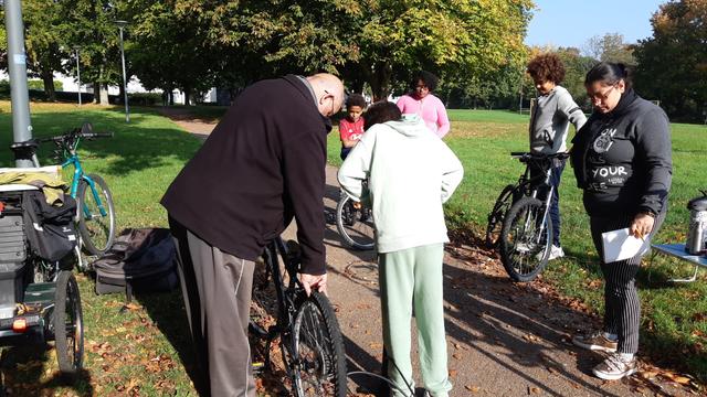 Homme aidant un jeune garçon à entretenir son vélo, entourés de 2 autres garçons à VTT et 2 jeunes femmes sur un grand pré arboré