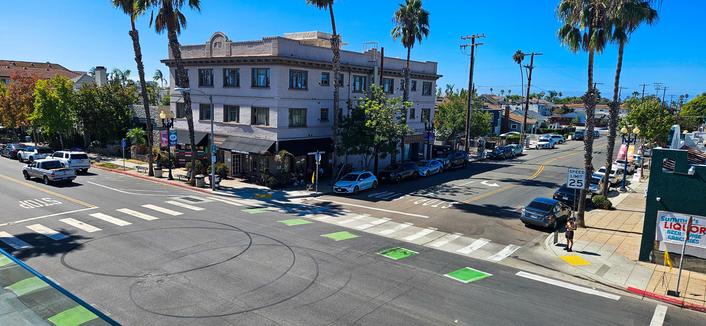 A wide-angle, daytime shot of a street intersection with a large, multi-story building as a focal point. The building is a light beige color with numerous windows and dark-colored storefronts on the ground level. Several vehicles are visible on the streets surrounding the intersection, and pedestrian crosswalks are marked with white stripes and green painted squares. Palm trees line the streets on both sides. A sign displaying “SPEED LIMIT 25” is visible on a pole near the intersection, and a sign on a building reads “Liquor.”