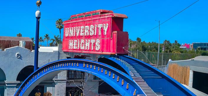 A brightly colored, arched sign stands in front of buildings on a sunny day. The sign is a large, red boxcar-shaped structure mounted on a curved, blue, tiled archway. The words “UNIVERSITY HEIGHTS” are illuminated in white neon lettering across the front of the boxcar. The archway has the word “BOULEVARD” repeated across the blue tiles. Palm trees and buildings are visible in the background, and a tall pole with a light fixture is positioned on the left side of the image.