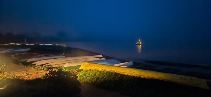 A nighttime view of a body of water with a series of illuminated structures along the shore. The shoreline features a terraced arrangement of white, curved shapes that are lit from below, creating a glowing effect. A wooden pier extends into the water, also illuminated, leading towards a small sailboat silhouetted against the dark water. The foreground is dark and appears to be covered in vegetation, with some light reflecting off of the ground near the structures. The sky is a deep, dark blue, and the water is calm, with a subtle reflection of the light from the structures and the boat.