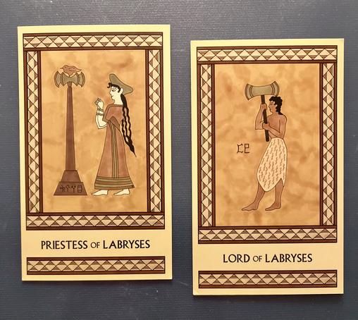 Two Minoan Tarot cards side by side on a dark gray surface. Both cards are painted in shades of gold, cream, and tan and have intricate borders. The Priestess of Labryses card, on the left, shows a woman with long hair, wearing an ankle-length robe and a hat. She is facing left, where a tall labrys stands with a bird perched on top of it. Her arms are bent at the elbows as she holds her clenched fists up in front of her in a sacred gesture. The Lord of Labryses card, on the right, shows a man wearing a knee-length animal hide skirt. He is shirtless and barefoot, with long hair. He faces left and holds up a large labrys in front of him.
