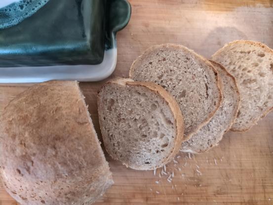 Ein sehr kleiner länglicher Laib Brot, zur Hälfte in Scheiben. Eine blau-weiße Butterschale in der linken oberen Ecke ist zum Teil zu sehen. Beides liegt auf einer Holzarbeitsplatte.