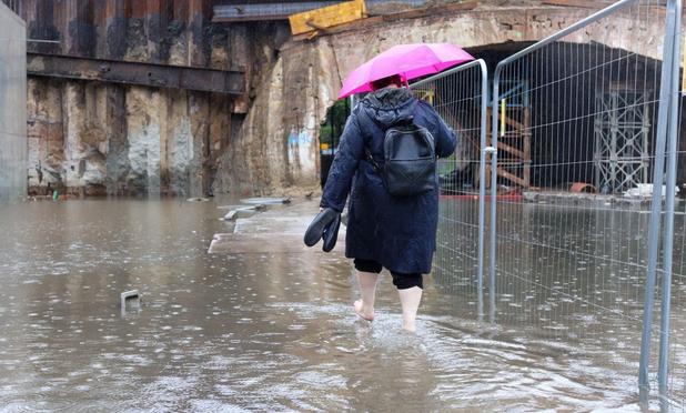 Mann von hinten mit Regenschirm wartet durch kniehohe Überschwemmung vor einer Unterführung, Rechter hand das Terrain durch bauzaun abgesperrt