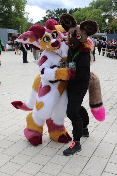 Two fursuiters posing outdoors. One is a hoofed dragon fullsuit, sporting white fur with brown and purple markings, some in the shape of hearts. The other is a hyena partial, with a nose ring and a pink-tipped tail. They have their arms around each other and are posing for the camera. In the background, people are milling about a food court.